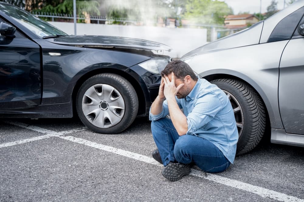 A distressed man sits on the ground in front of a car accident.