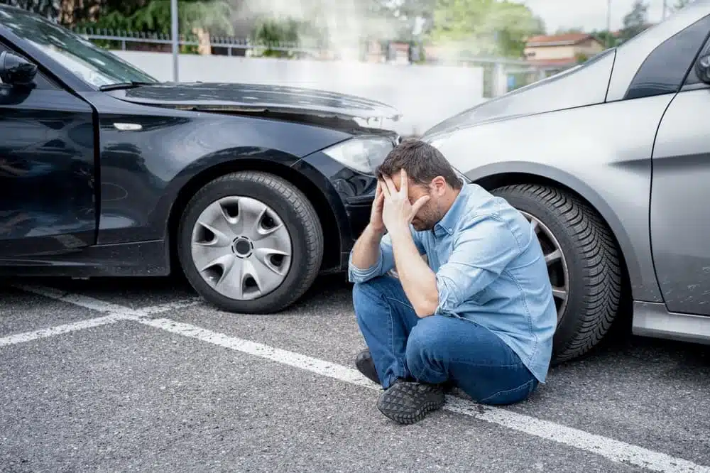 A distressed man sits on the ground in front of a car accident.