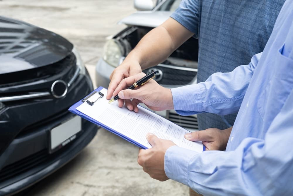 Two people review a document on a clipboard in front of damaged cars.